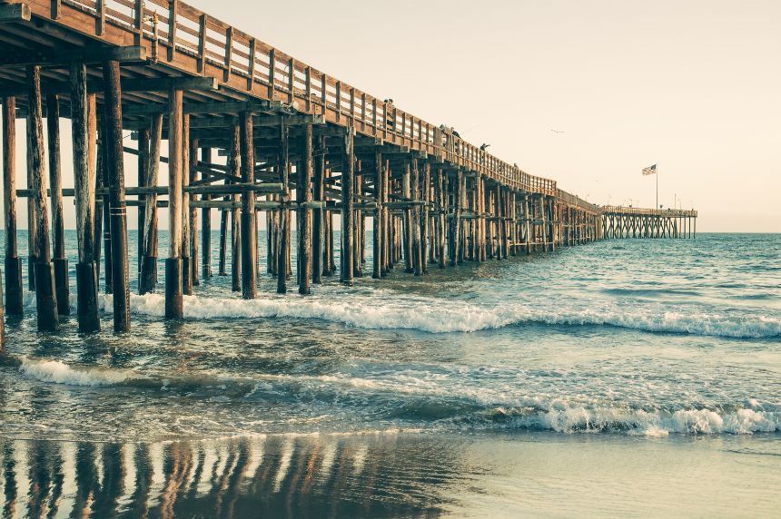 A long wooden pier stretches into the ocean with waves gently crashing below and an American flag flying at the end.