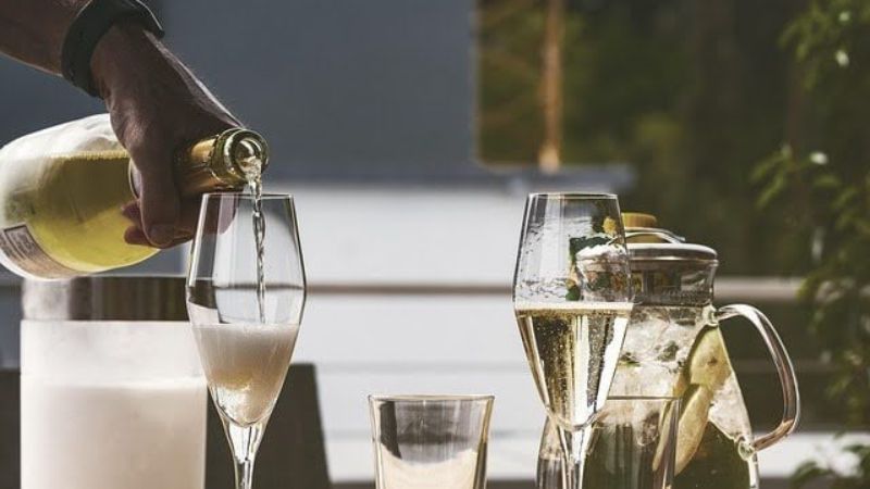 Hand pouring champagne into a glass beside a pitcher of lemon water.